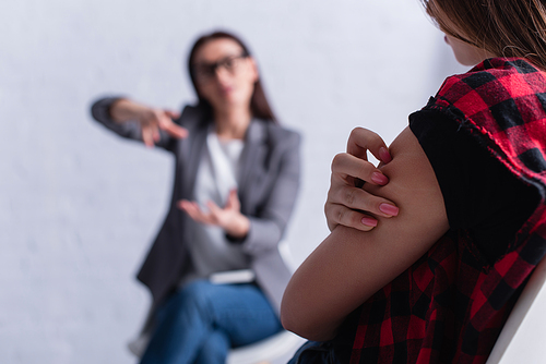 teenage patient scratching skin near psychologist on blurred foreground