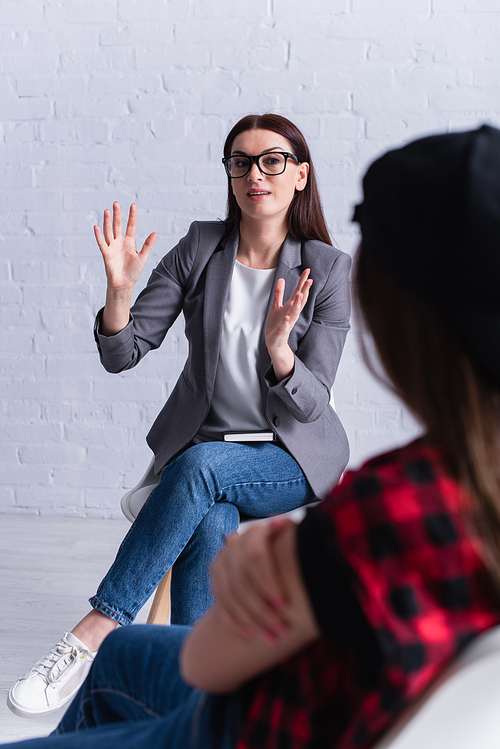 psychologist in glasses gesturing while looking at teenager on blurred foreground