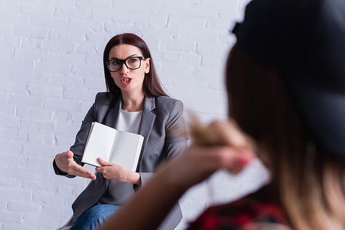 psychologist in glasses holding blank notebook and gesturing while looking at teenage patient on blurred foreground