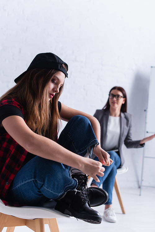 depressed teenage girl in cap sitting near psychologist on blurred background