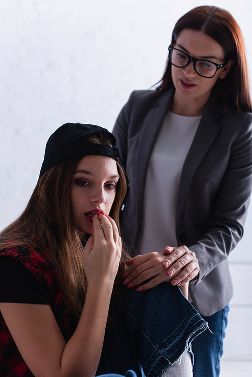 worried teenage girl in cap biting nails near psychologist on blurred background