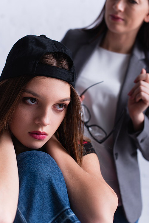 displeased teenage girl in cap near psychologist on blurred background