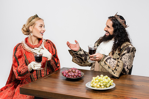 happy interracial historical couple in crowns holding glasses of red wine while sitting at table with grapes isolated on white