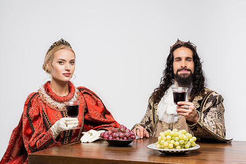 interracial historical couple in crowns holding glasses of red wine while sitting at table with grapes isolated on white