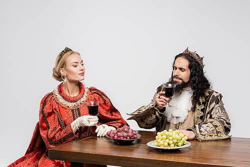 blonde queen looking at hispanic king in medieval clothing smelling red wine in glass isolated on white