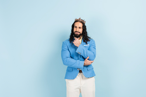 pensive and bearded hispanic man in crown and jacket posing on blue