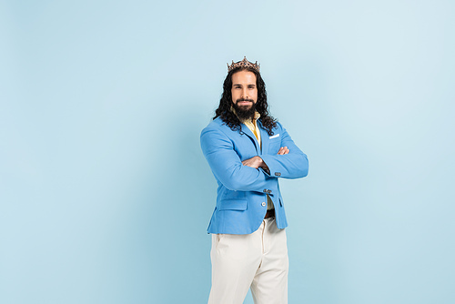 bearded hispanic man in crown and jacket standing with crossed arms on blue