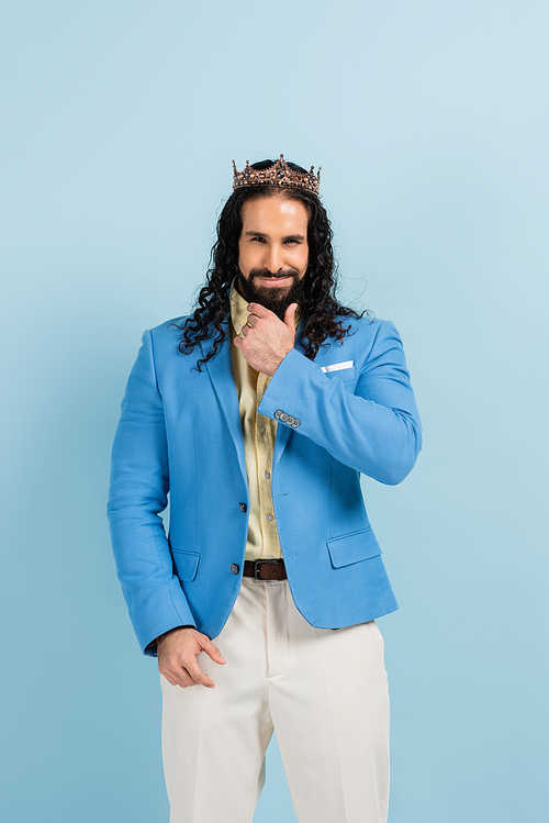 bearded hispanic man in crown and jacket smiling isolated on blue
