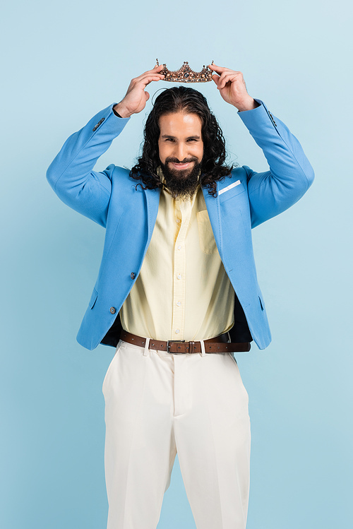 bearded hispanic man in jacket wearing crown and smiling isolated on blue