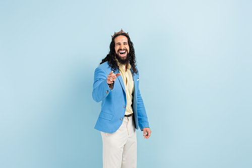 bearded hispanic man in crown and jacket laughing while pointing with finger on blue