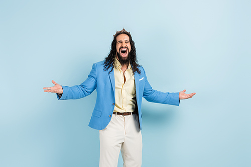 excited hispanic man in crown and jacket standing with outstretched hands on blue
