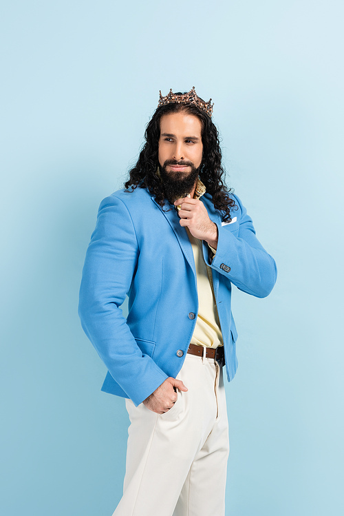 bearded hispanic man in crown and jacket posing with hand in pocket isolated on blue