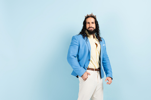 bearded hispanic man in crown and jacket standing with hand in pocket on blue