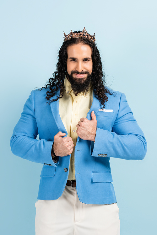 cheerful hispanic man in crown adjusting jacket isolated on blue
