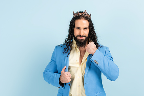 bearded hispanic man in crown adjusting jacket isolated on blue