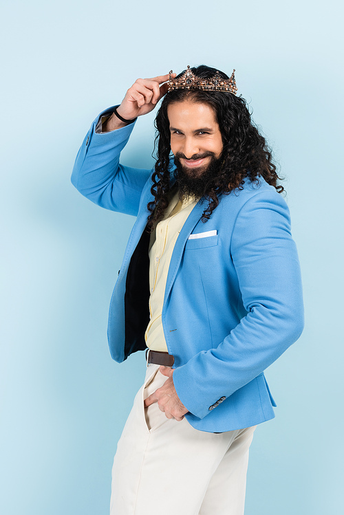 smiling and curly hispanic man in jacket adjusting golden crown isolated on blue