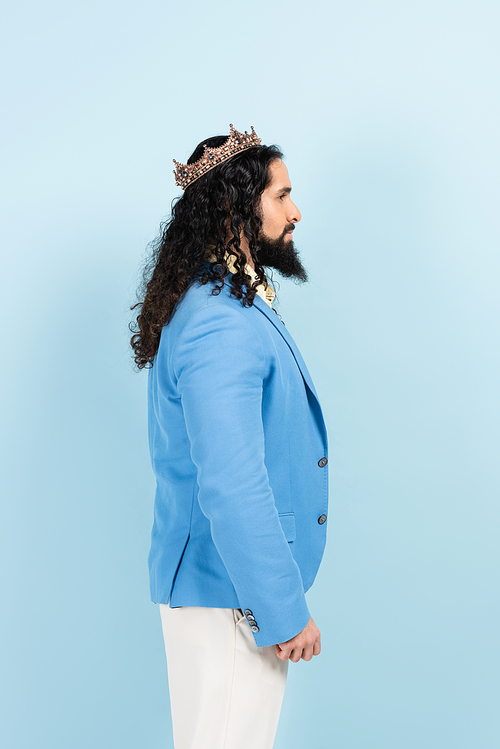 side view of bearded hispanic man in jacket and crown isolated on blue