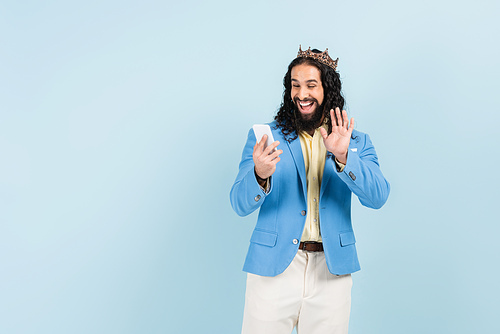 bearded hispanic man in jacket and crown waving hand during video call isolated on blue