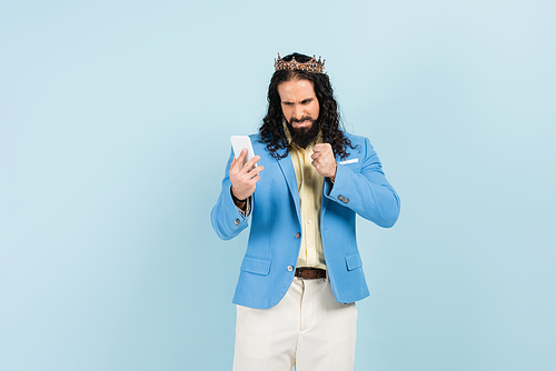 angry hispanic man in jacket and crown showing clenched fist and holding smartphone isolated on blue