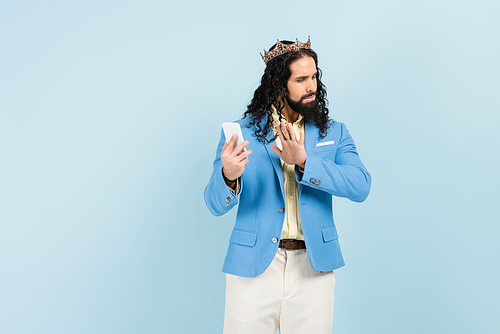 offended hispanic man in jacket and crown showing stop gesture while holding smartphone isolated on blue