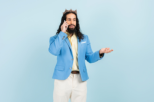 bearded hispanic man in jacket and crown talking on smartphone isolated on blue