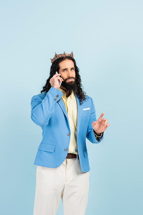 bearded hispanic man in jacket and crown talking on cellphone isolated on blue