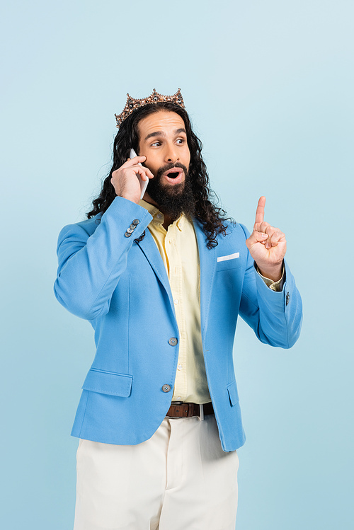 surprised hispanic man in jacket and crown talking on smartphone isolated on blue