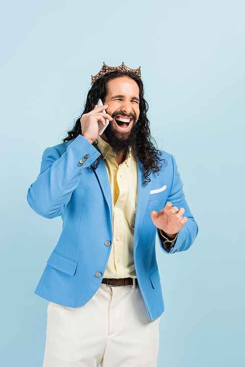 happy hispanic man in jacket and crown talking on smartphone and laughing isolated on blue