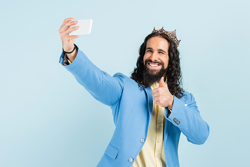 happy hispanic man in jacket and crown showing thumb up and taking selfie isolated on blue