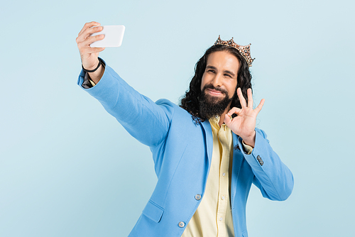 happy hispanic man in jacket and crown showing okay and taking selfie isolated on blue