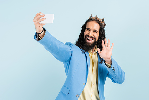 happy hispanic man in jacket and crown holding smartphone and having video call isolated on blue
