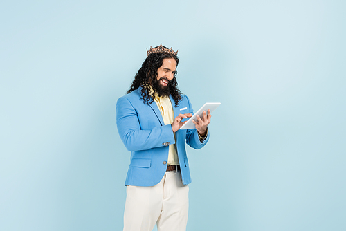 happy hispanic man in jacket and crown holding digital tablet isolated on blue