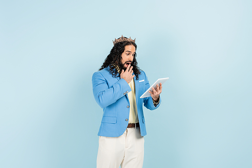 shocked hispanic man in jacket and crown holding digital tablet isolated on blue