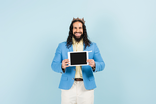 happy hispanic man in jacket and crown holding digital tablet with blank screen isolated on blue
