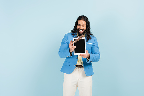 happy hispanic man in jacket holding digital tablet with blank screen and crown isolated on blue