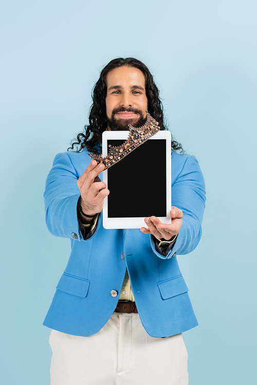 cheerful hispanic man in jacket holding digital tablet with blank screen and crown isolated on blue
