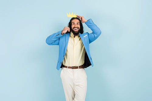 happy hispanic man in jacket holding paper crown on stick isolated on blue