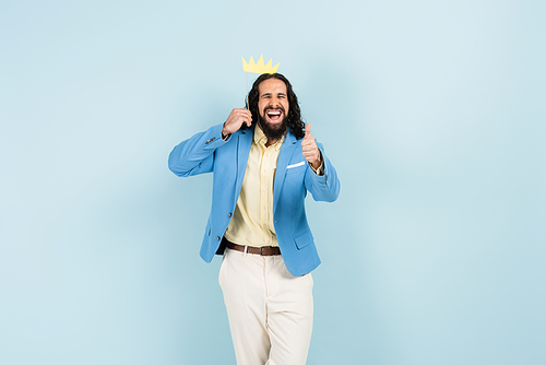 happy hispanic man in jacket holding paper crown on stick and showing thumb up isolated on blue