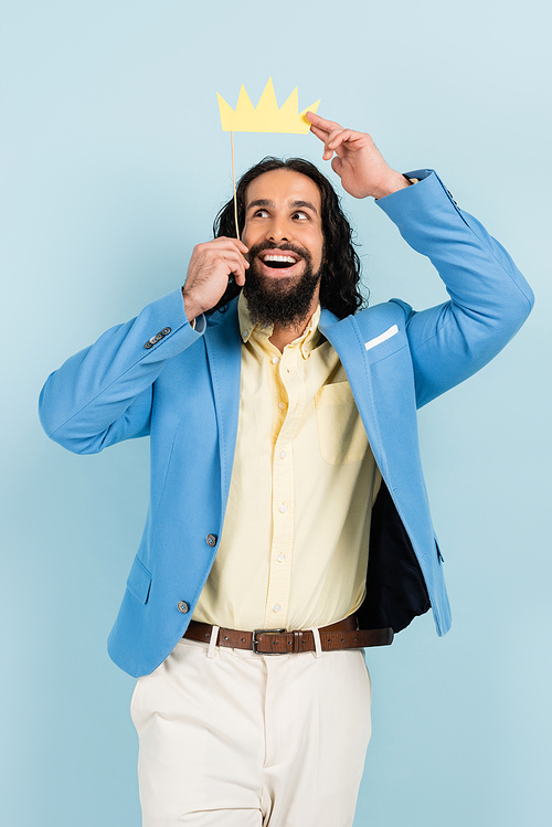 joyful hispanic man in jacket holding paper crown on stick isolated on blue