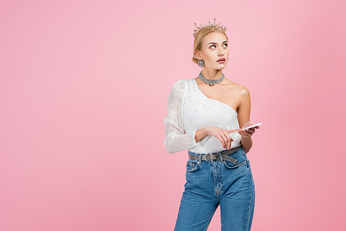 pensive blonde woman in luxury crown standing with smartphone isolated on pink