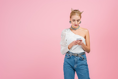 blonde woman in luxury crown using smartphone isolated on pink