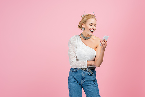 cheerful woman in luxury crown holding smartphone isolated on pink