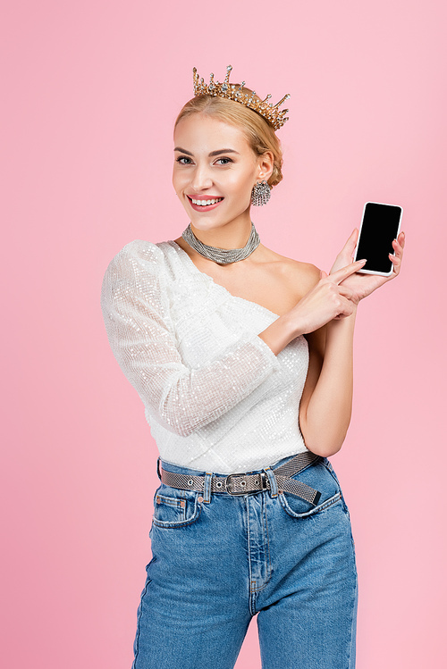 happy blonde woman in luxury crown holding smartphone with blank screen isolated on pink