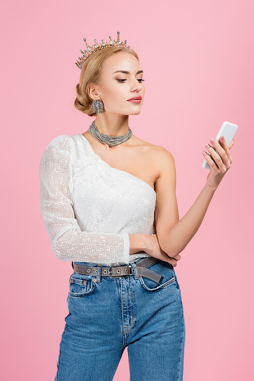 blonde woman in luxury crown looking at smartphone isolated on pink