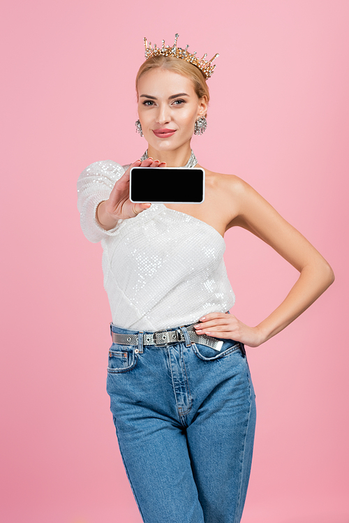 happy blonde woman in crown holding smartphone with blank screen isolated on pink