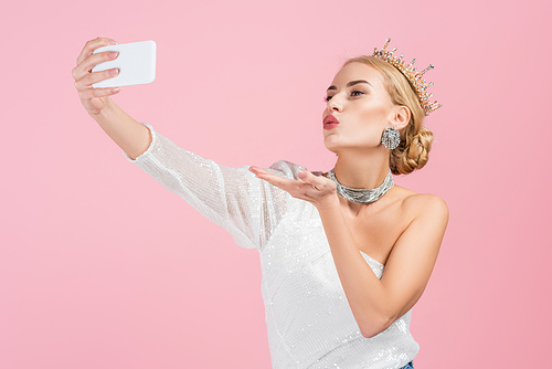blonde woman in luxury crown taking selfie on smartphone and sending air kiss isolated on pink
