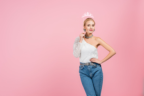 stylish woman holding paper crown on stick and standing with hand on hip isolated on pink