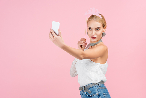 blonde woman holding paper crown on stick and taking selfie on smartphone isolated on pink