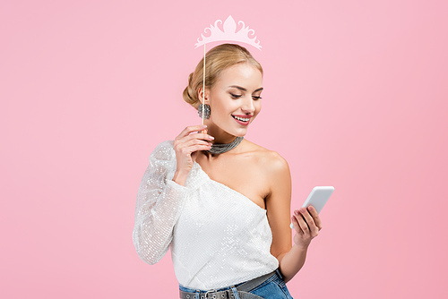 happy woman holding paper crown on stick and smartphone isolated on pink