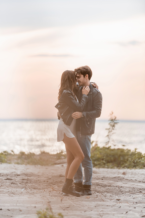 Selective focus of young man embracing girlfriend in dress and leather jacket on beach at sunset